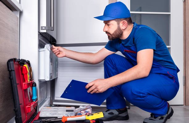 Technician crouching beside a refrigerator with a clipboard and tool box